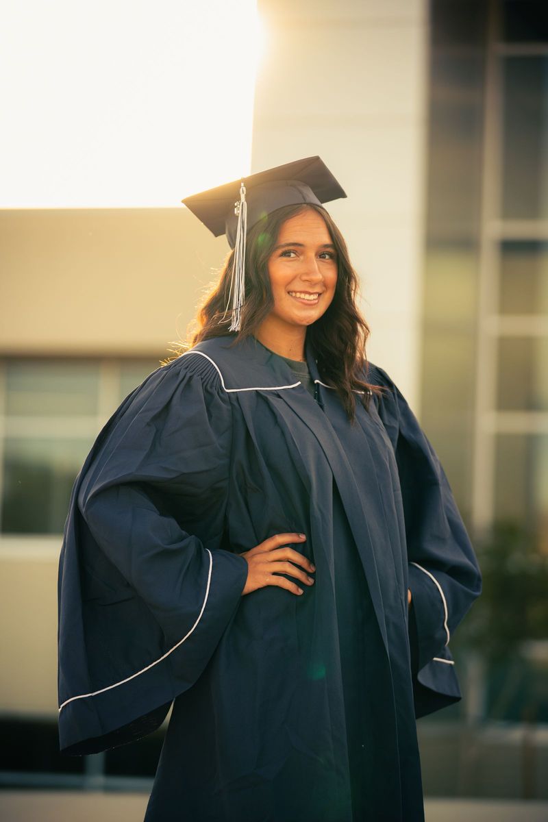 Graduate in cap and gown with a hand on her hip, low sun behind modern buildings