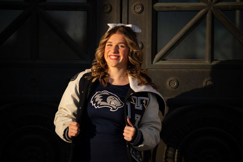 Cheerleader smiling in a letterman jacket against dark bronze doors, warm side light