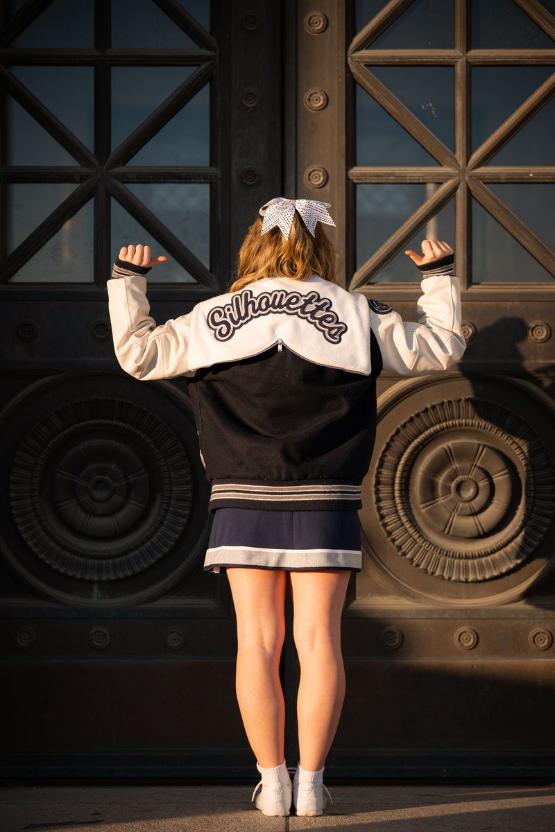 Cheerleader seen from behind, arms raised in a lettered jacket before ornate bronze doors