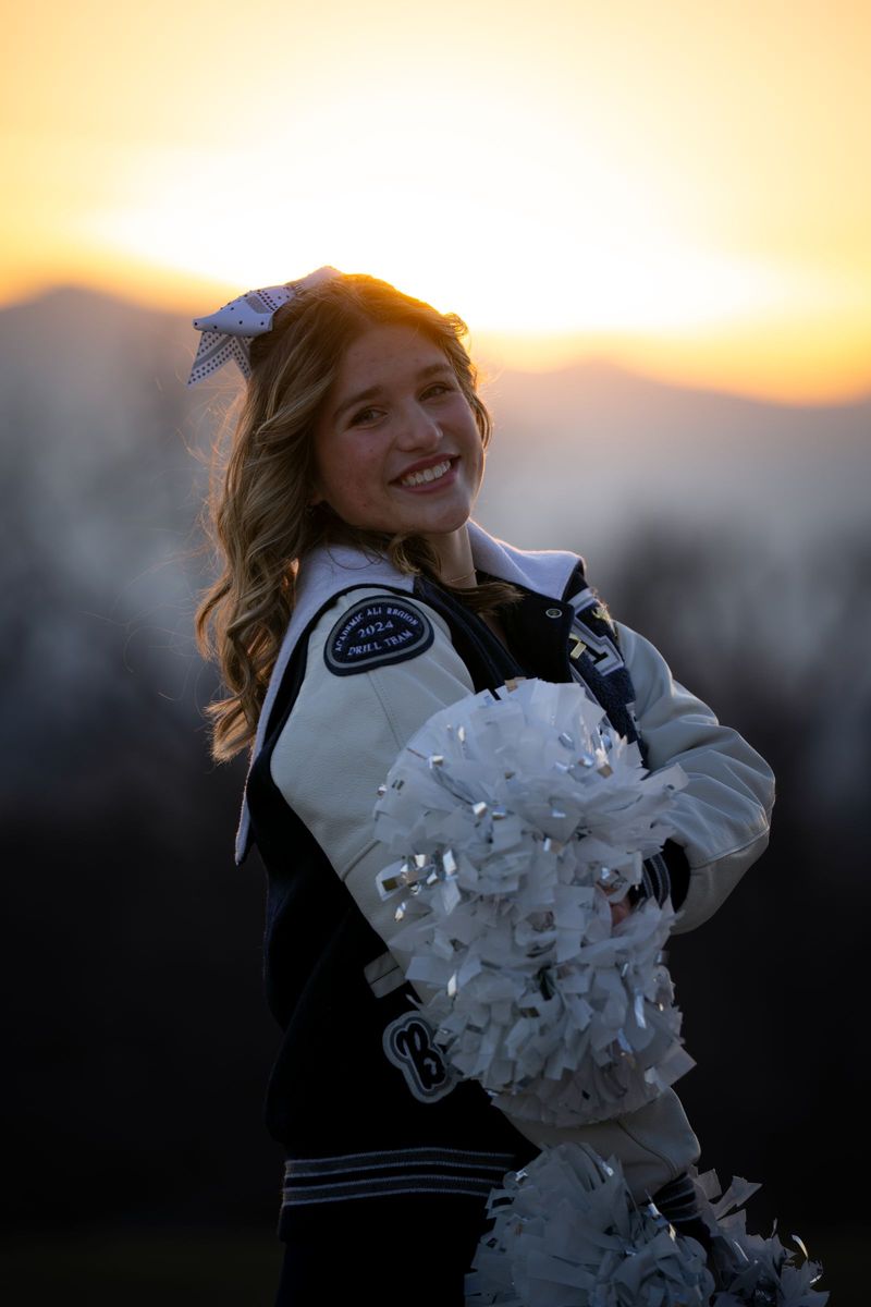 Smiling cheerleader in a letterman jacket holding pom-poms, backlit by sunset over mountains