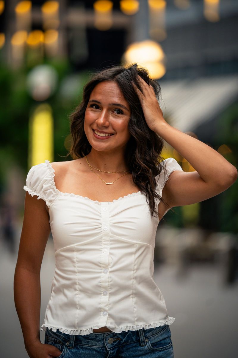 Senior in a white top with a hand in her hair on a blurred city street