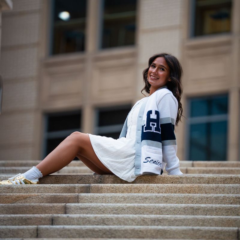 Senior in a white dress and letterman sweater seated on wide stone steps