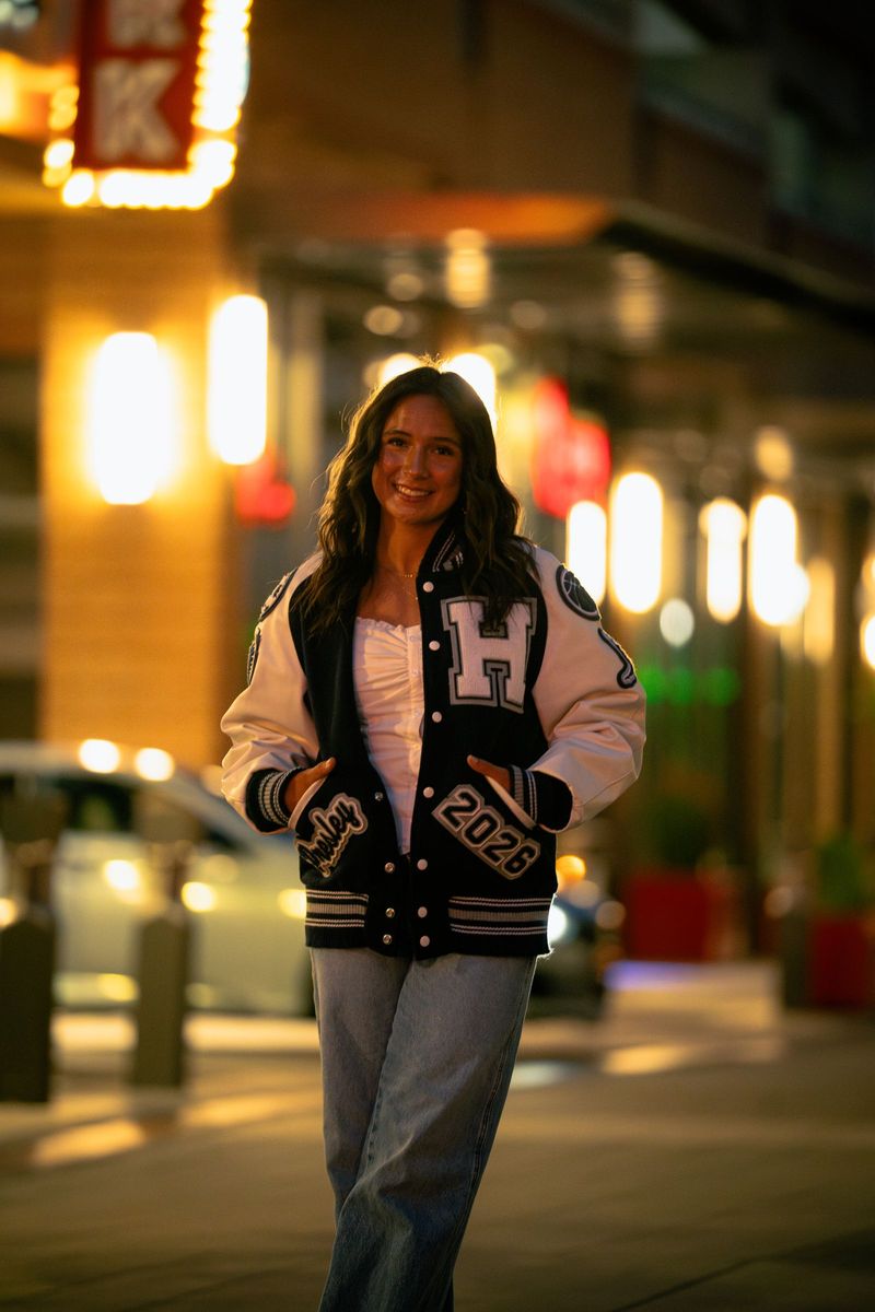 Senior in a letterman jacket on a city street at dusk, glowing signs behind