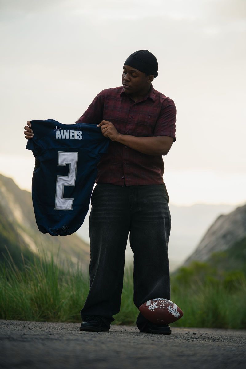 Senior holding up a football jersey with a football at his feet, mountain canyon behind
